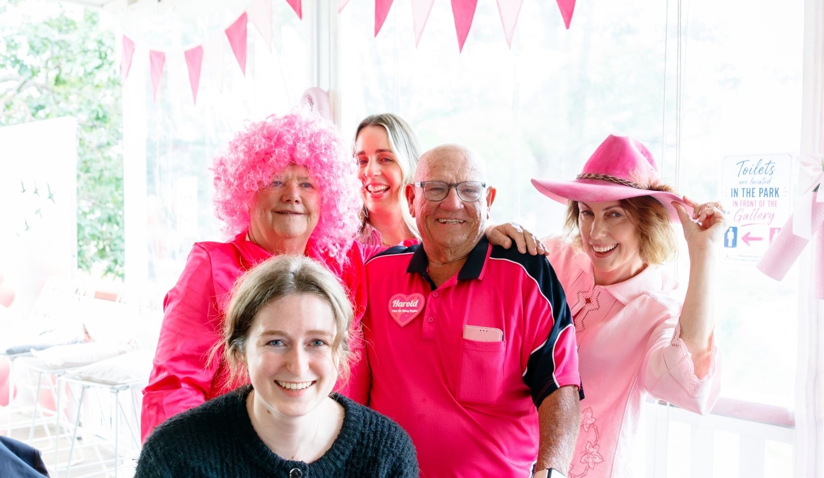 Harold and four other Pink Up Kilcoy supporters pose together indoors, dressed in shades of pink and standing beneath pink bunting at a community fundraising event.