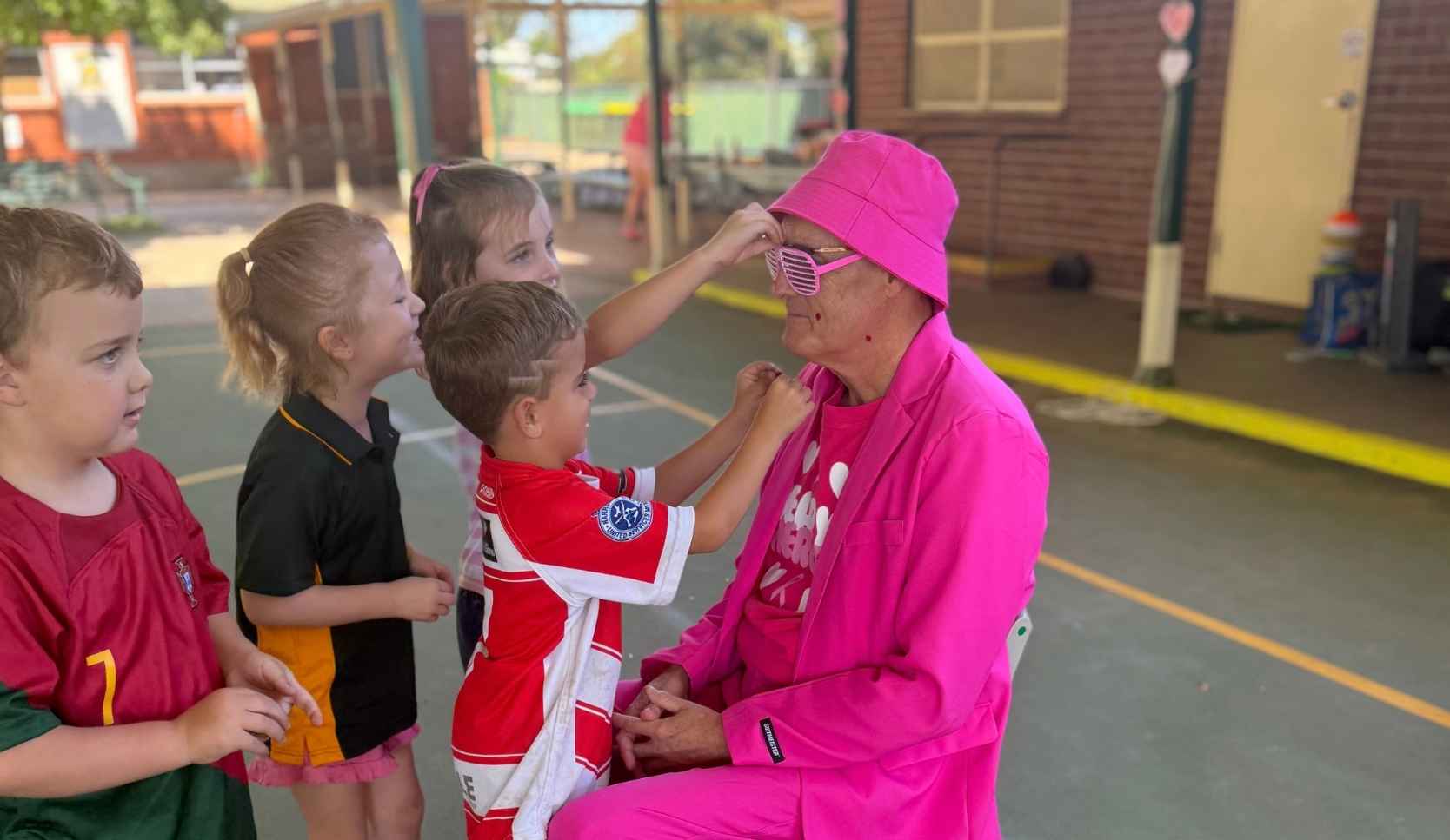 Man in a bright pink suit crouching outdoors while young children adjust his pink sunglasses and bucket hat
