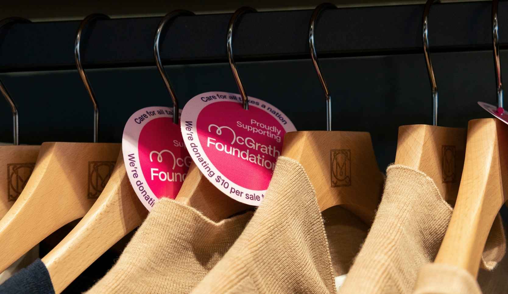 Two pink heart-shaped tags reading "Proudly Supporting McGrath Foundation" attached to wooden hangers holding beige jumpers