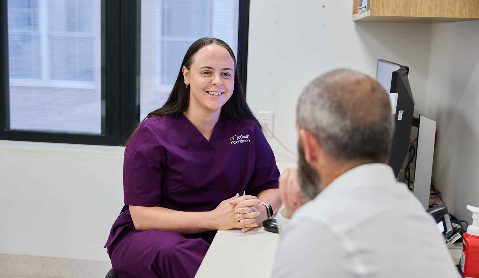 Woman in purple McGrath Foundation scrubs smiling while talking with a man in a white shirt at a desk