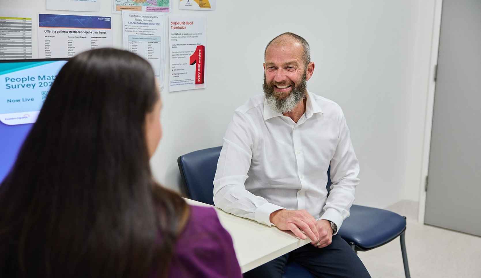 Bearded man in a white shirt smiling while seated across a table from a woman in purple scrubs in a hospital consulting room
