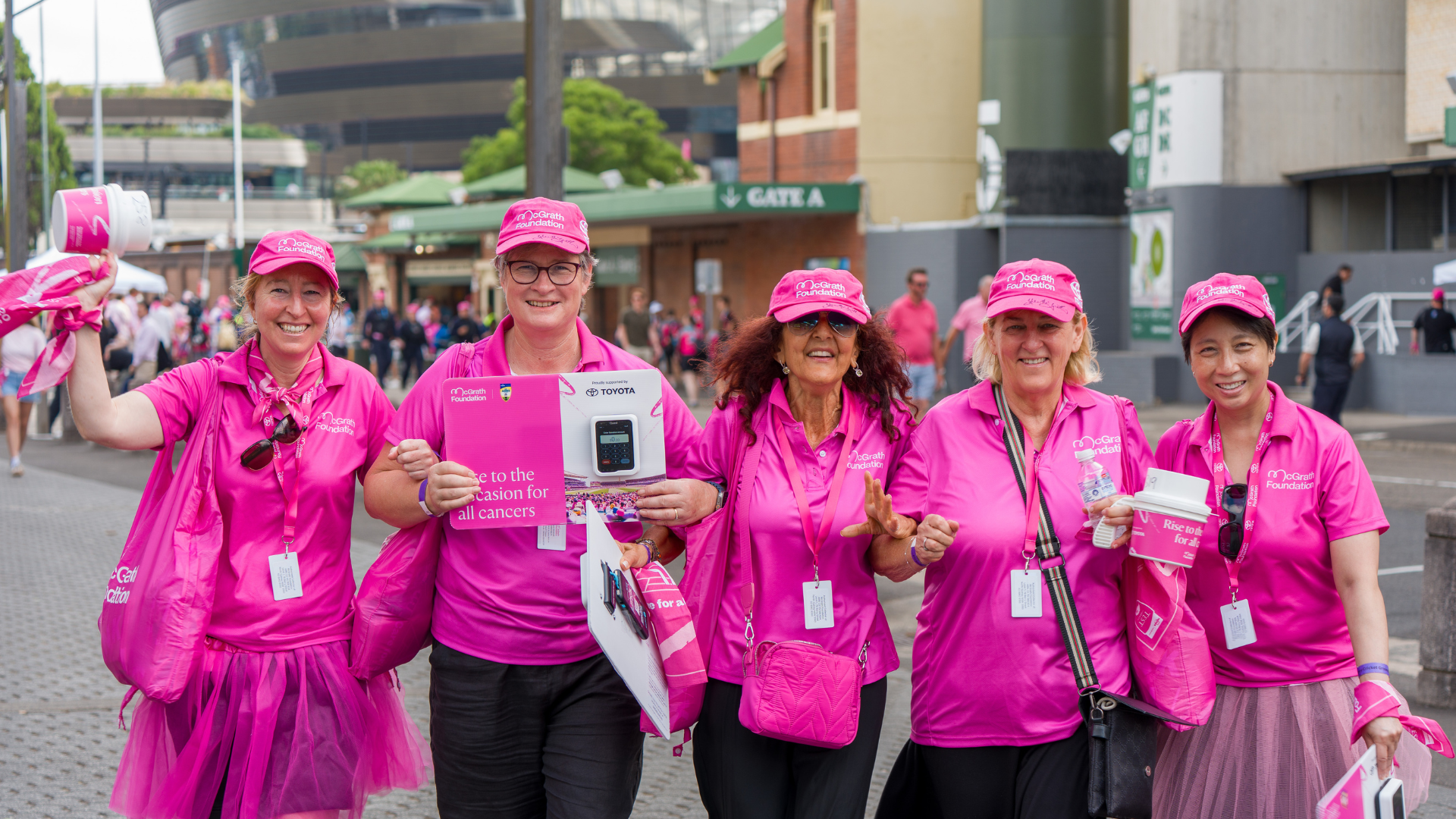 Five people in pink McGrath Foundation shirts and caps walk together outside the SCG, holding promotional items