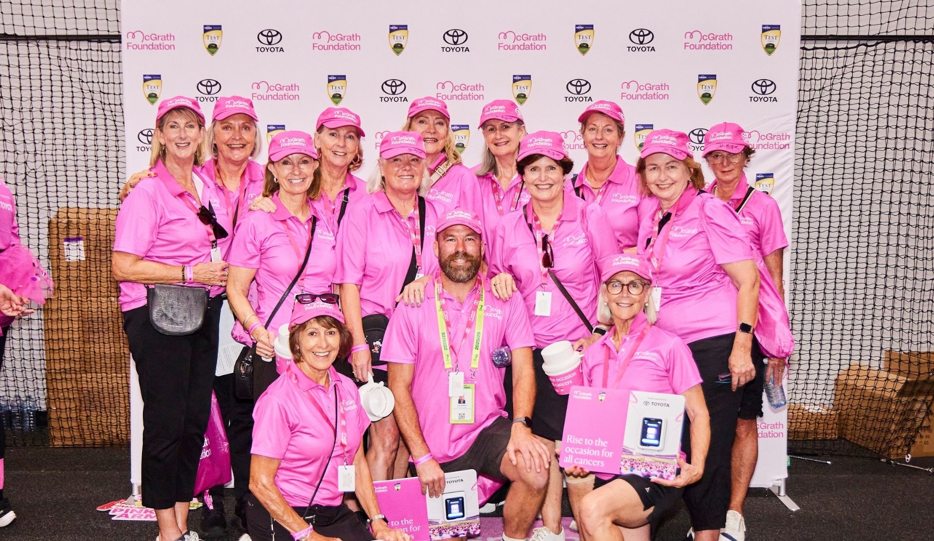 Group of McGrath Foundation volunteers in pink polos and caps posing at a Pink Test event, several kneeling with signs reading "Rise to the occasion for all cancers.