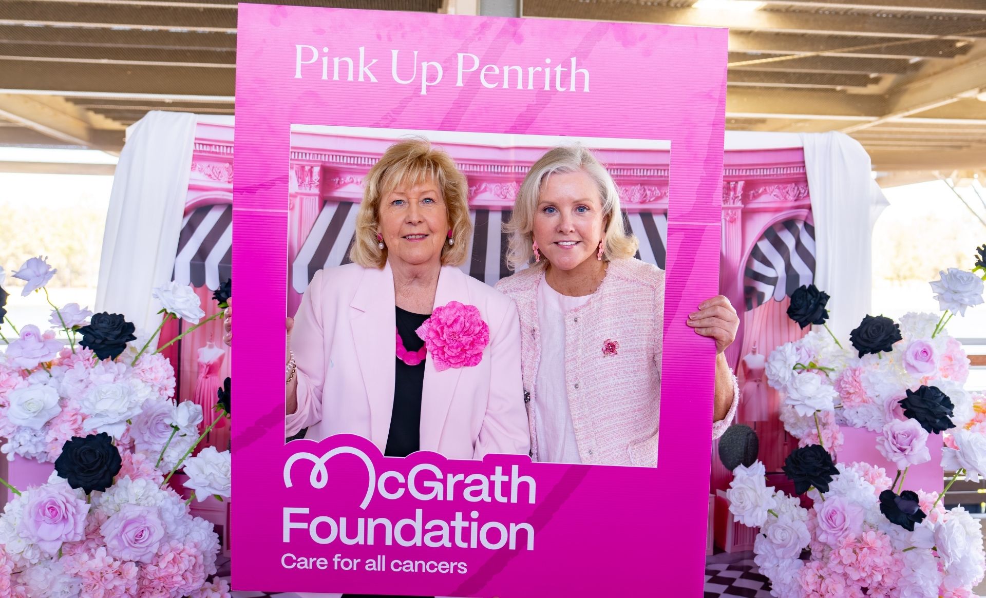 A fundraiser poses with Tracy Bevan inside a large pink “Pink Up Penrith” photo frame surrounded by pink, white and black floral decorations.