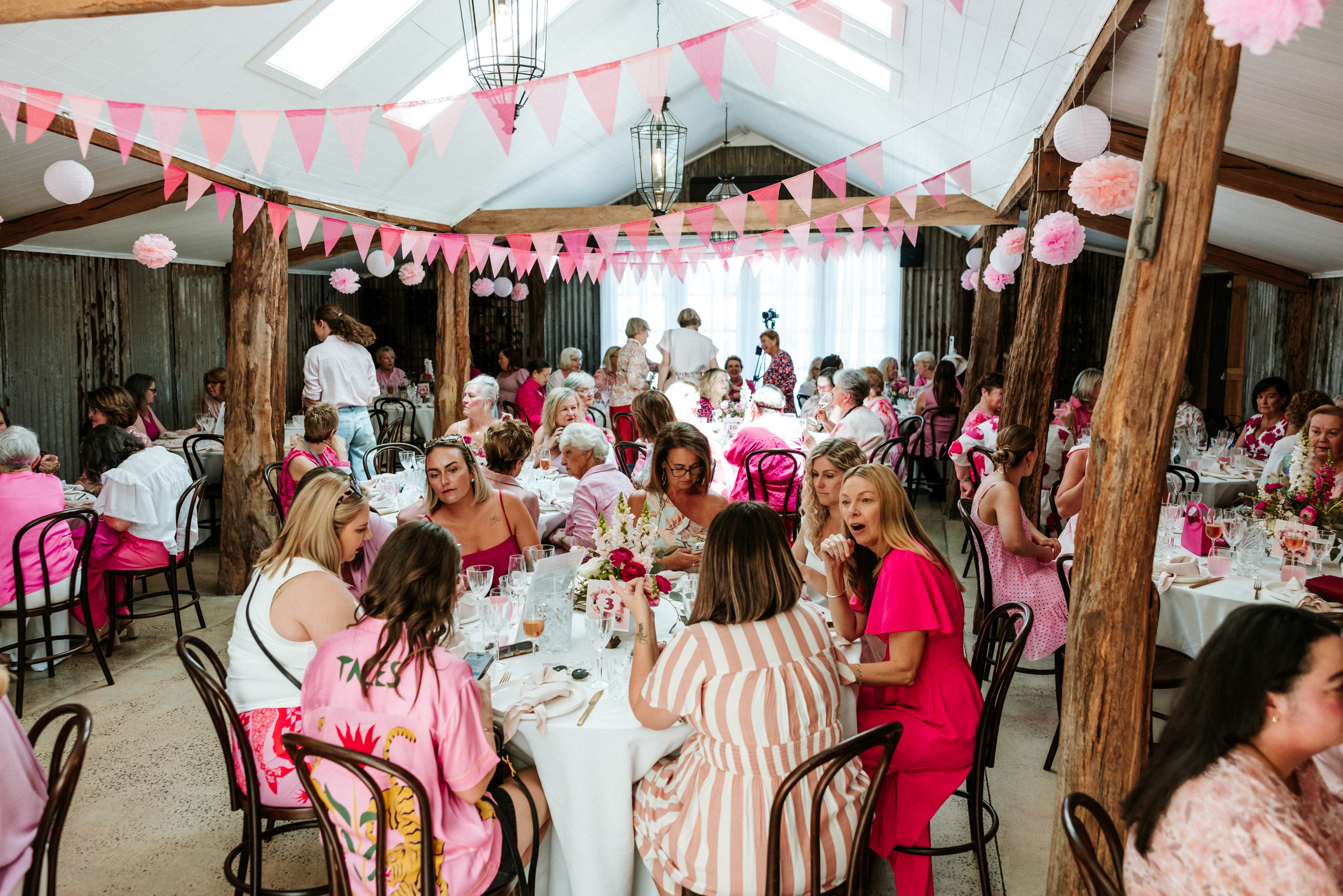 A large indoor event features tables of people dressed in pink seated under timber beams and pink bunting decorations, gathered for a McGrath Foundation-themed function.