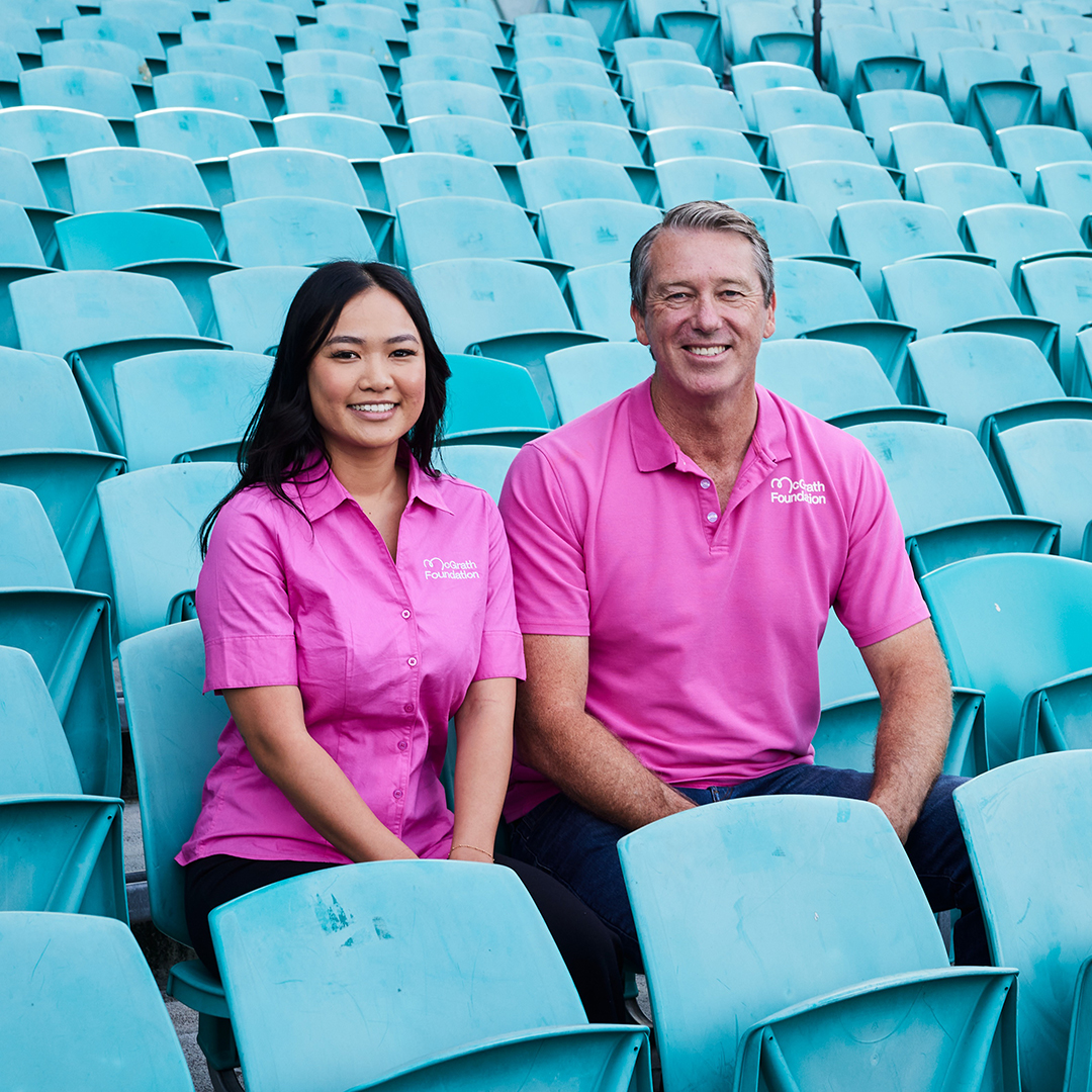 Man and woman wearing pink McGrath Foundation shirts sitting in blue seats of a cricket stadium.