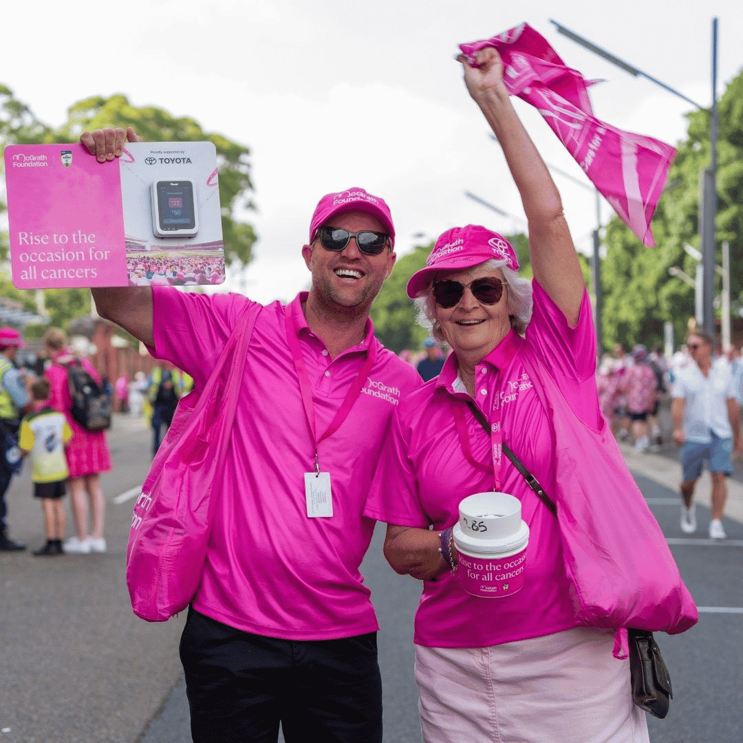 Two volunteers in bright pink McGrath Foundation clothing hold a fundraising sign and wave a pink scarf