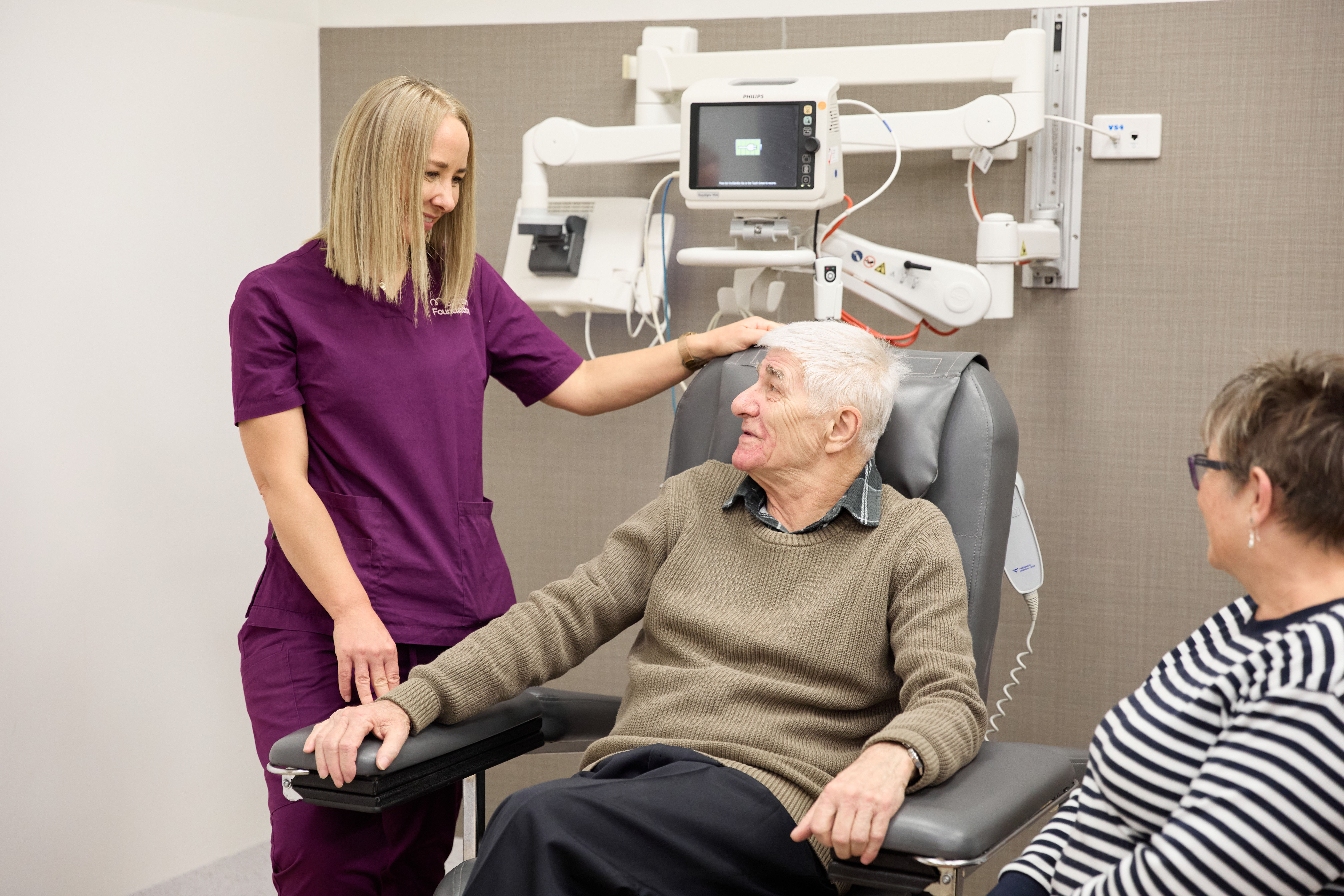 Man in chair connected to a medical machine talking to a McGrath Foundation nurse. Another woman sits in separate chair to the right.