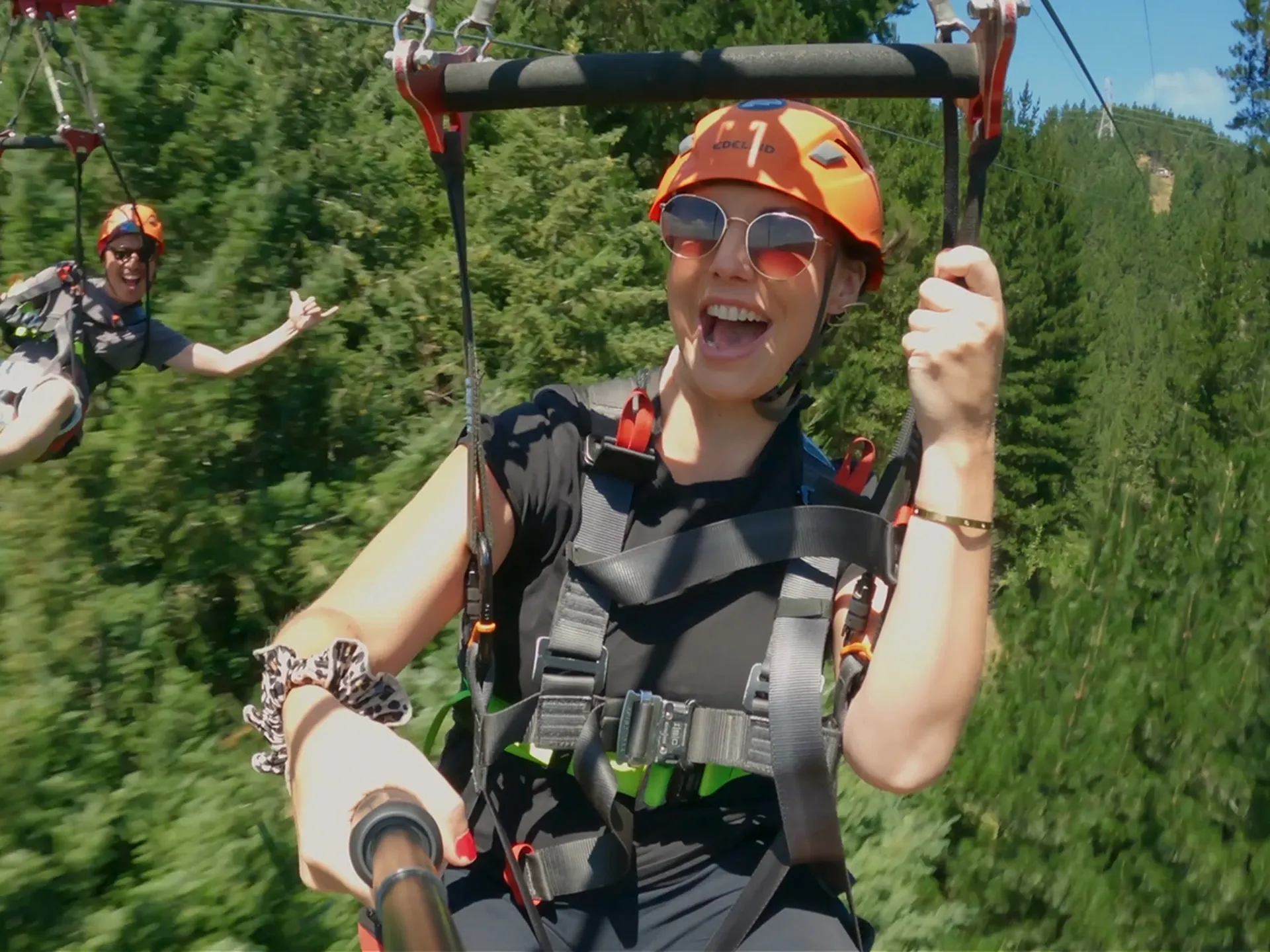 Two people wearing helmets and harnesses zip-line through a lush forest, smiling and enjoying the adventure on a sunny day.