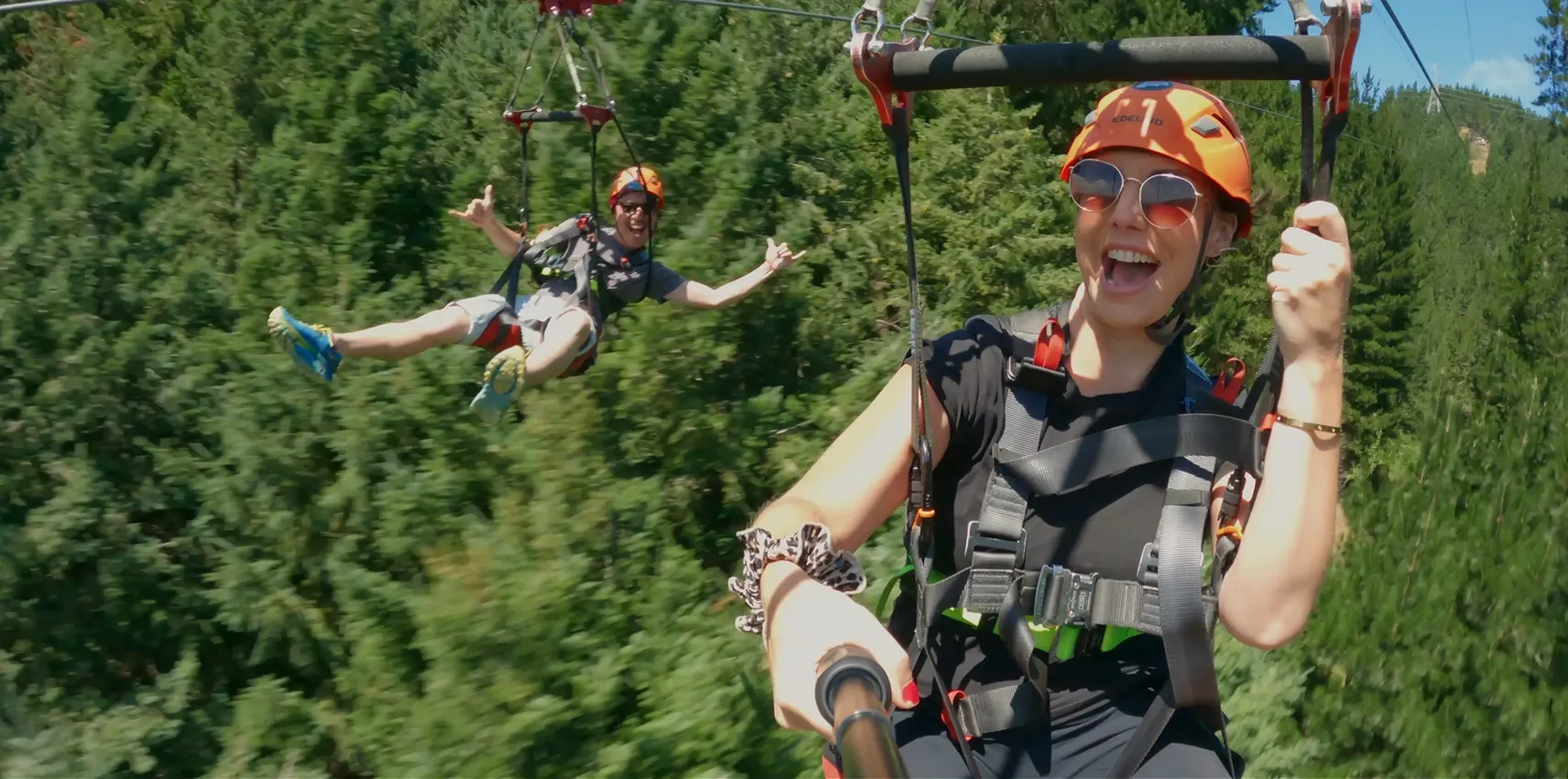Two people wearing helmets and harnesses zip-line through a lush forest, smiling and enjoying the adventure on a sunny day.