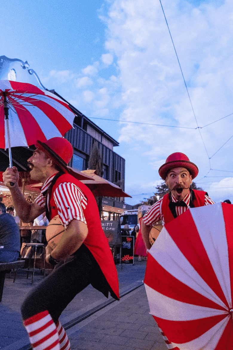 Two performers in red outfits with striped umbrellas entertain people at an outdoor venue during twilight.