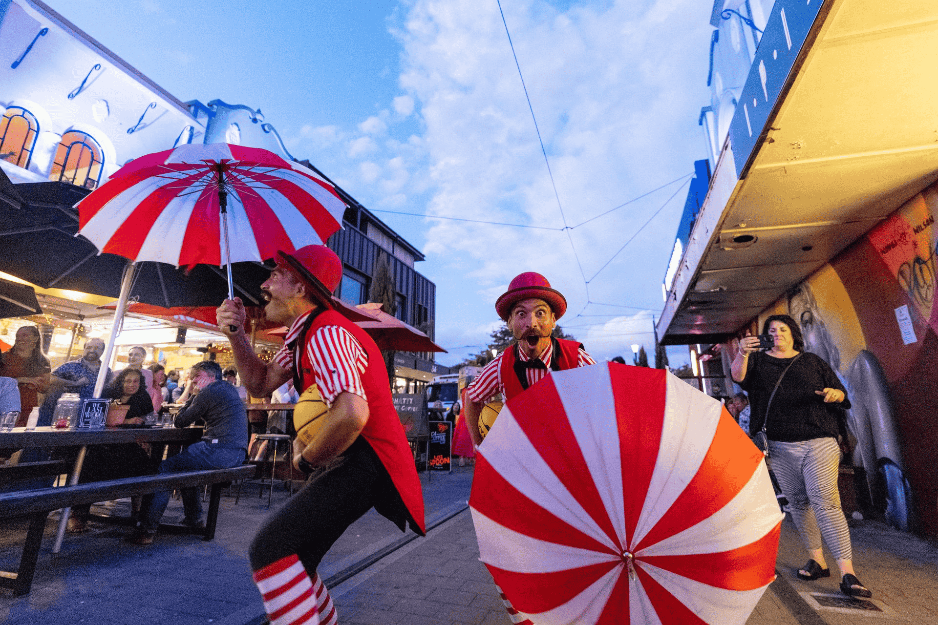 Two performers in red outfits with striped umbrellas entertain people at an outdoor venue during twilight.