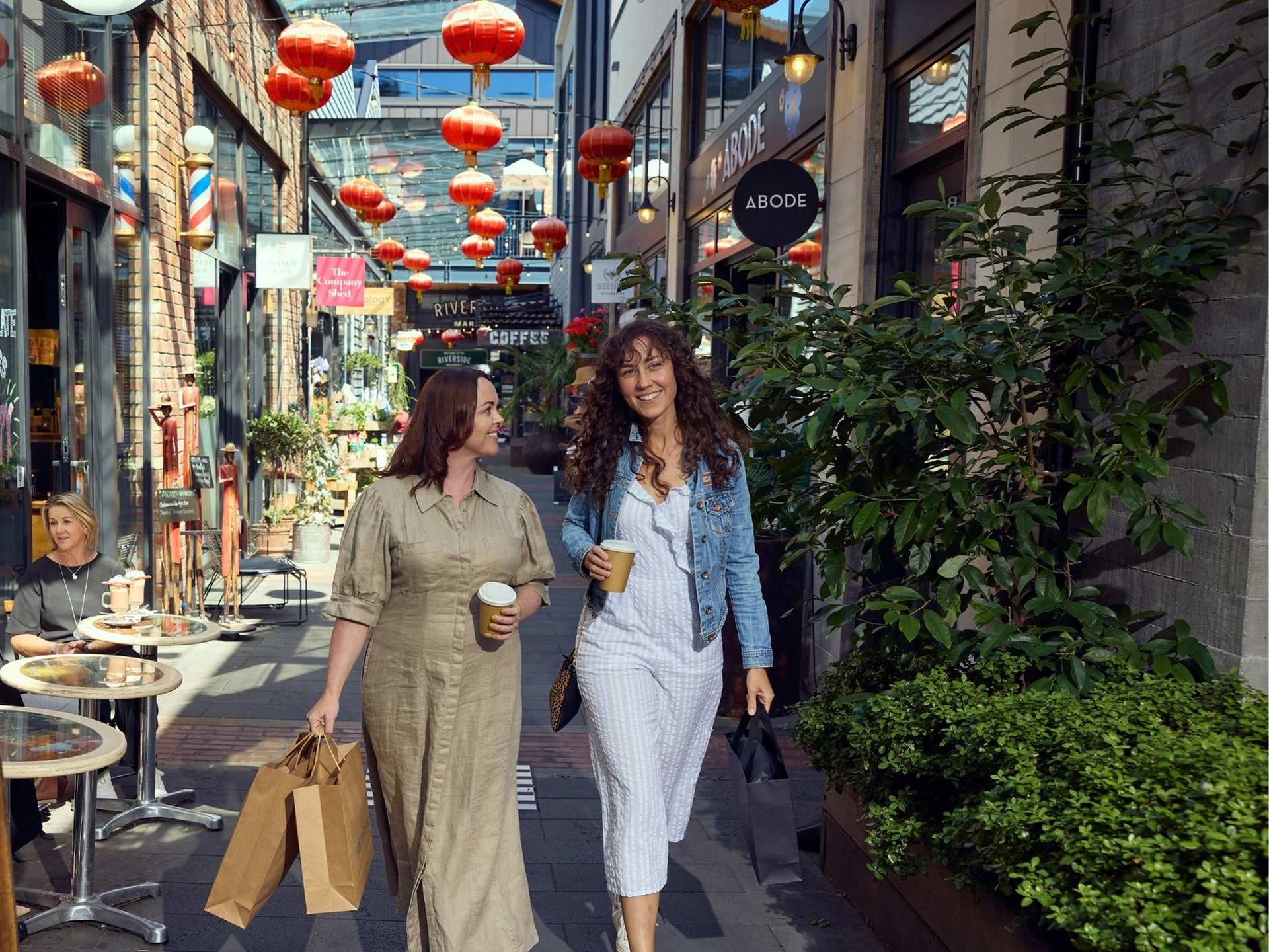 Two women walk down a lively, lantern-decorated alley, holding coffee cups and shopping bags, with people seated at outdoor café tables.