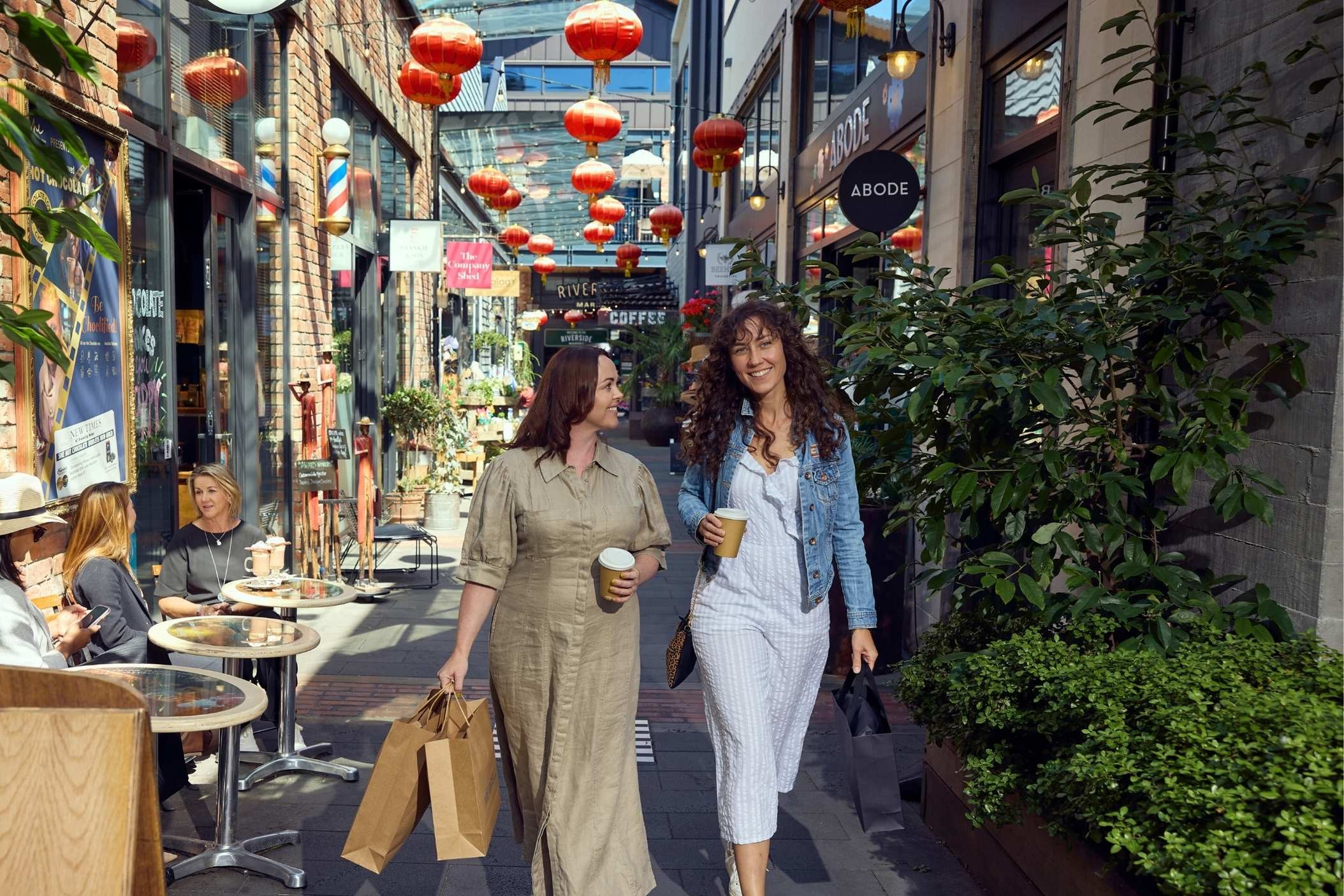 Two women walk down a lively, lantern-decorated alley, holding coffee cups and shopping bags, with people seated at outdoor café tables.