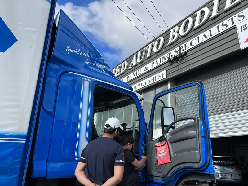 Two panel beaters working on a blue truck cab outside the Allied Auto Bodies workshop