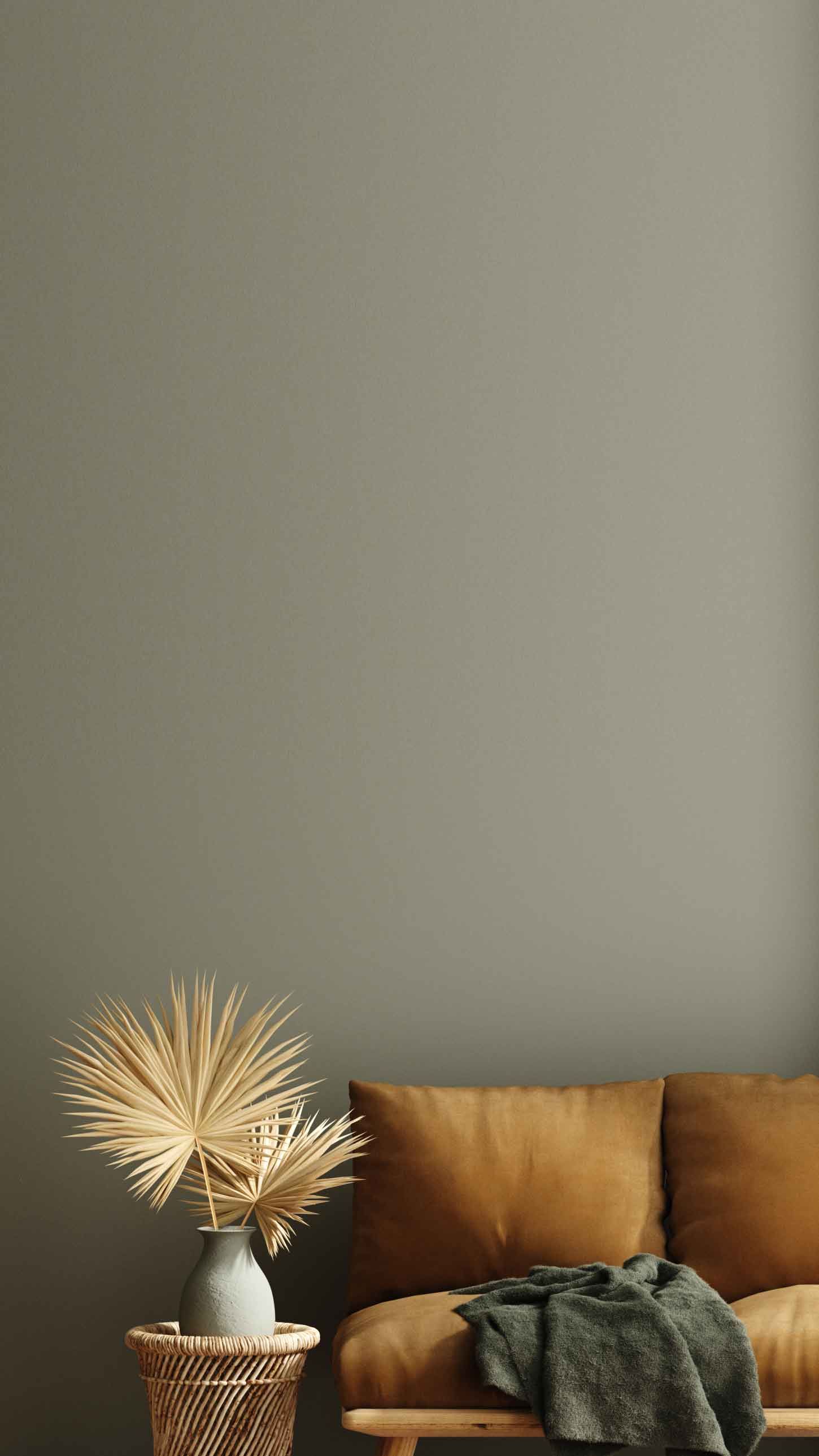 A minimalist living room with a brown sofa, a gray vase with dried palm leaves on a wicker table, and a dark gray wall in the background.