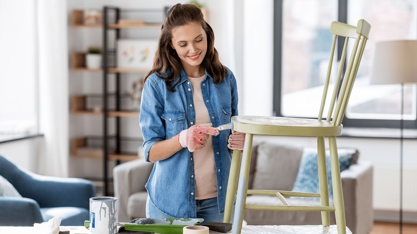 Woman painting a wooden chair green in a bright living room, wearing a denim shirt and gloves, with painting supplies on the table.