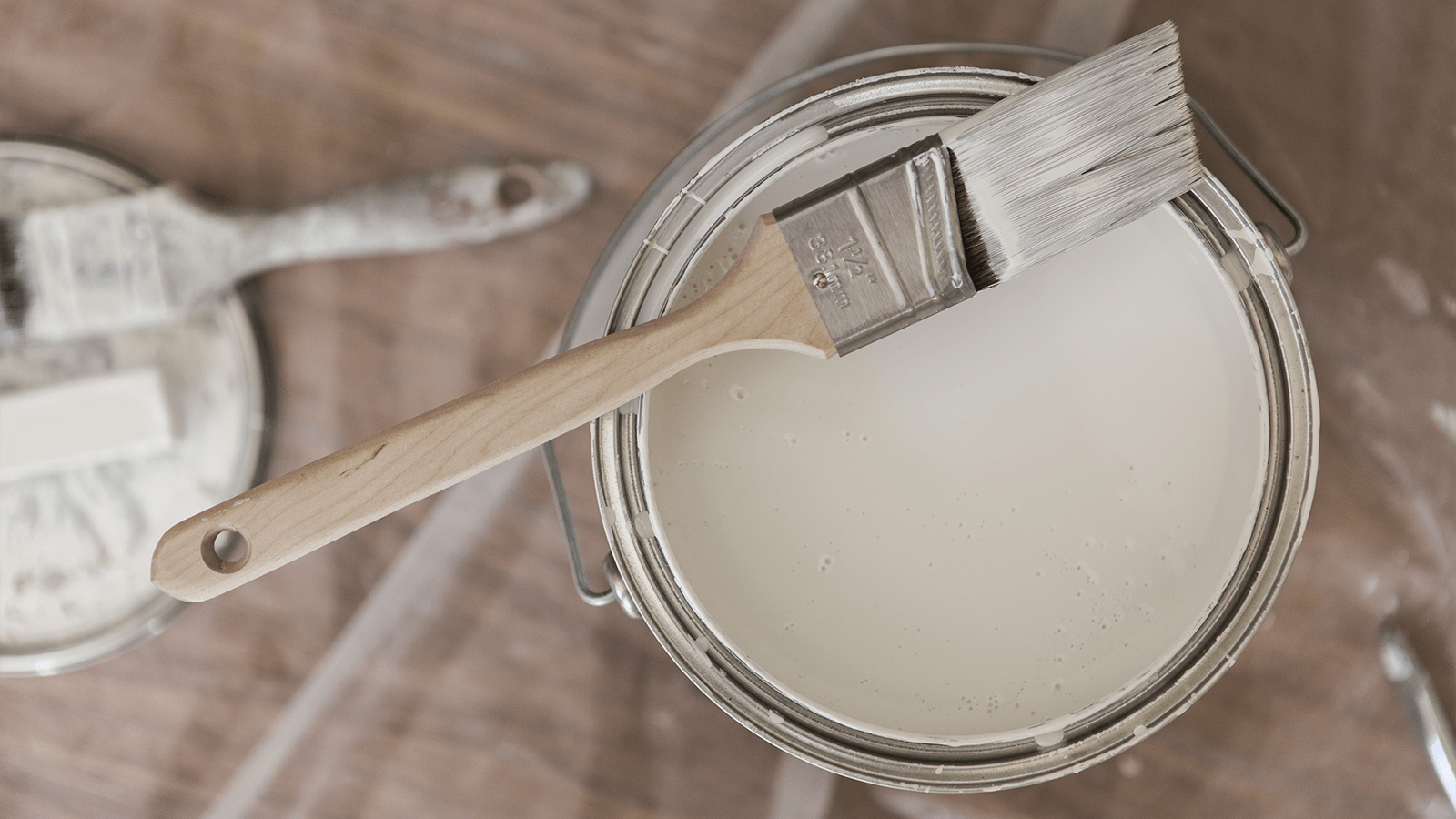 Open paint can with white paint, a paintbrush resting on top, and another brush nearby on a wooden surface.