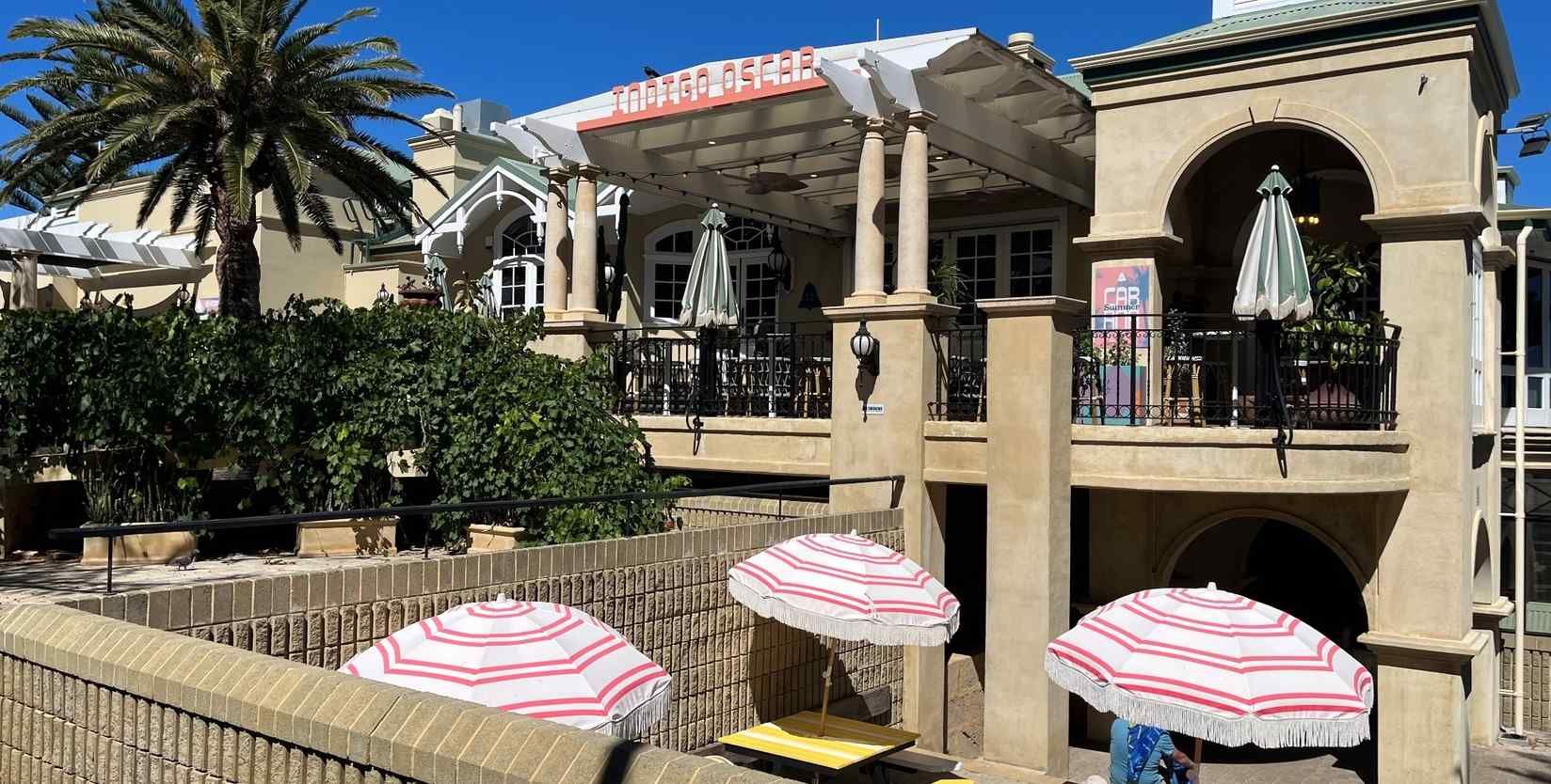 Outdoor cafe with red-striped umbrellas, arched windows, and palm tree. Sunny day with clear blue sky.