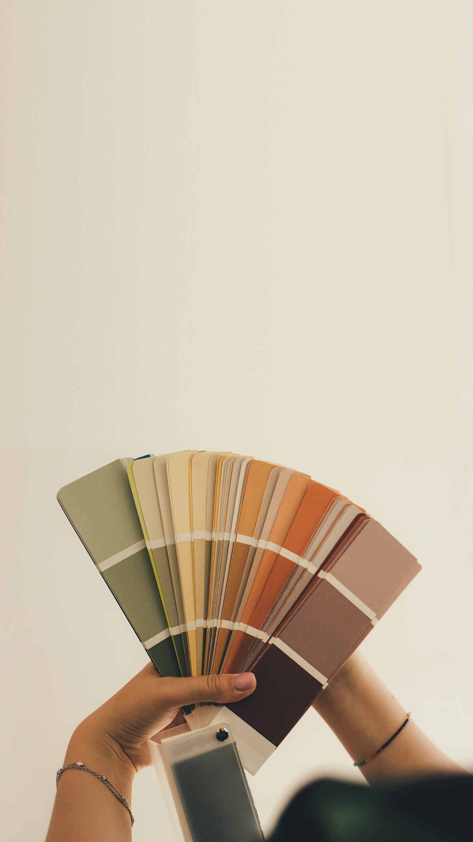 A person holding a fan of paint color swatches, featuring shades of green, yellow, orange, and brown, against a neutral background.