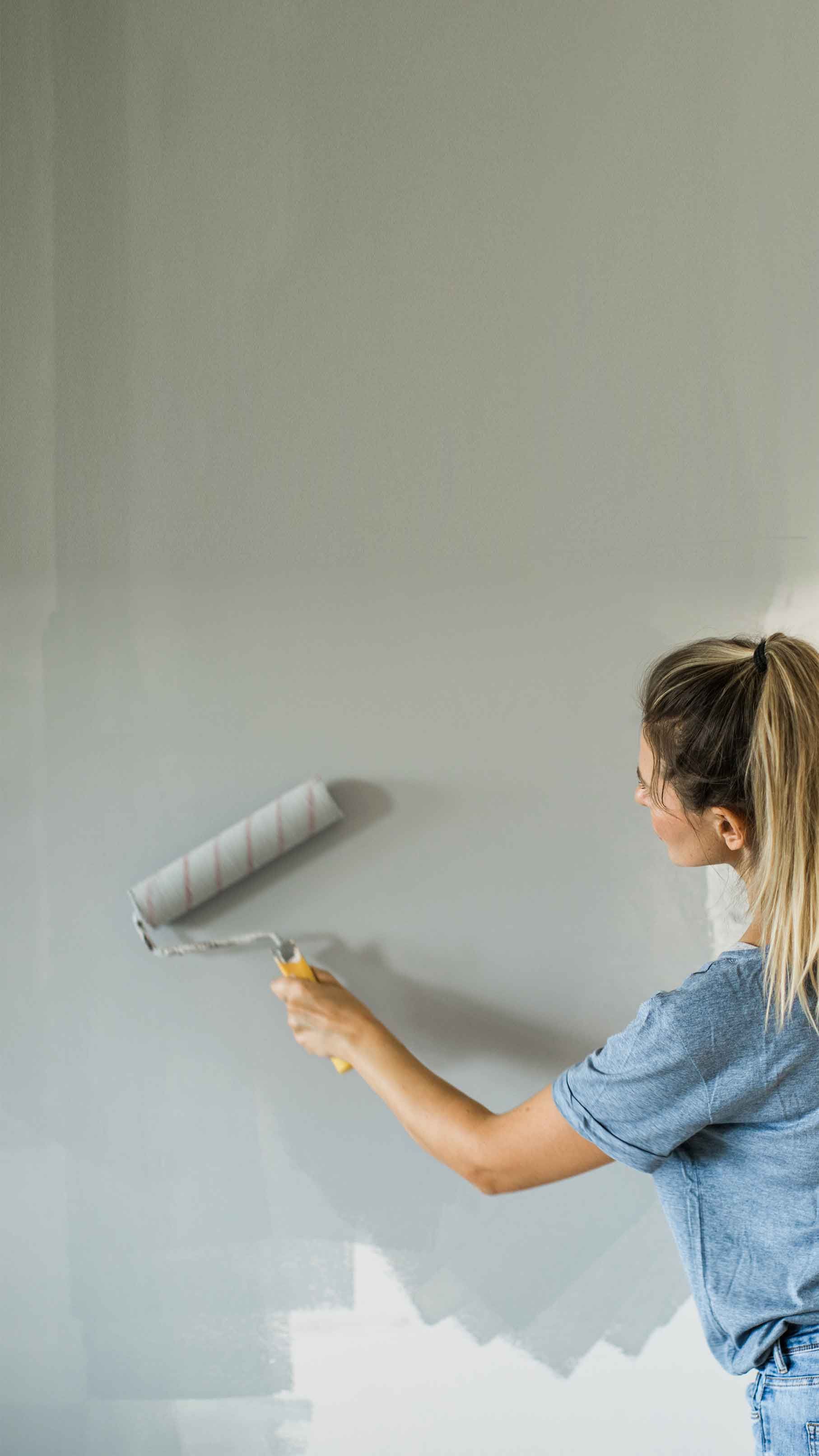 Woman painting a wall light gray with a roller. She wears a blue shirt and has her hair in a ponytail. The wall is partially painted.
