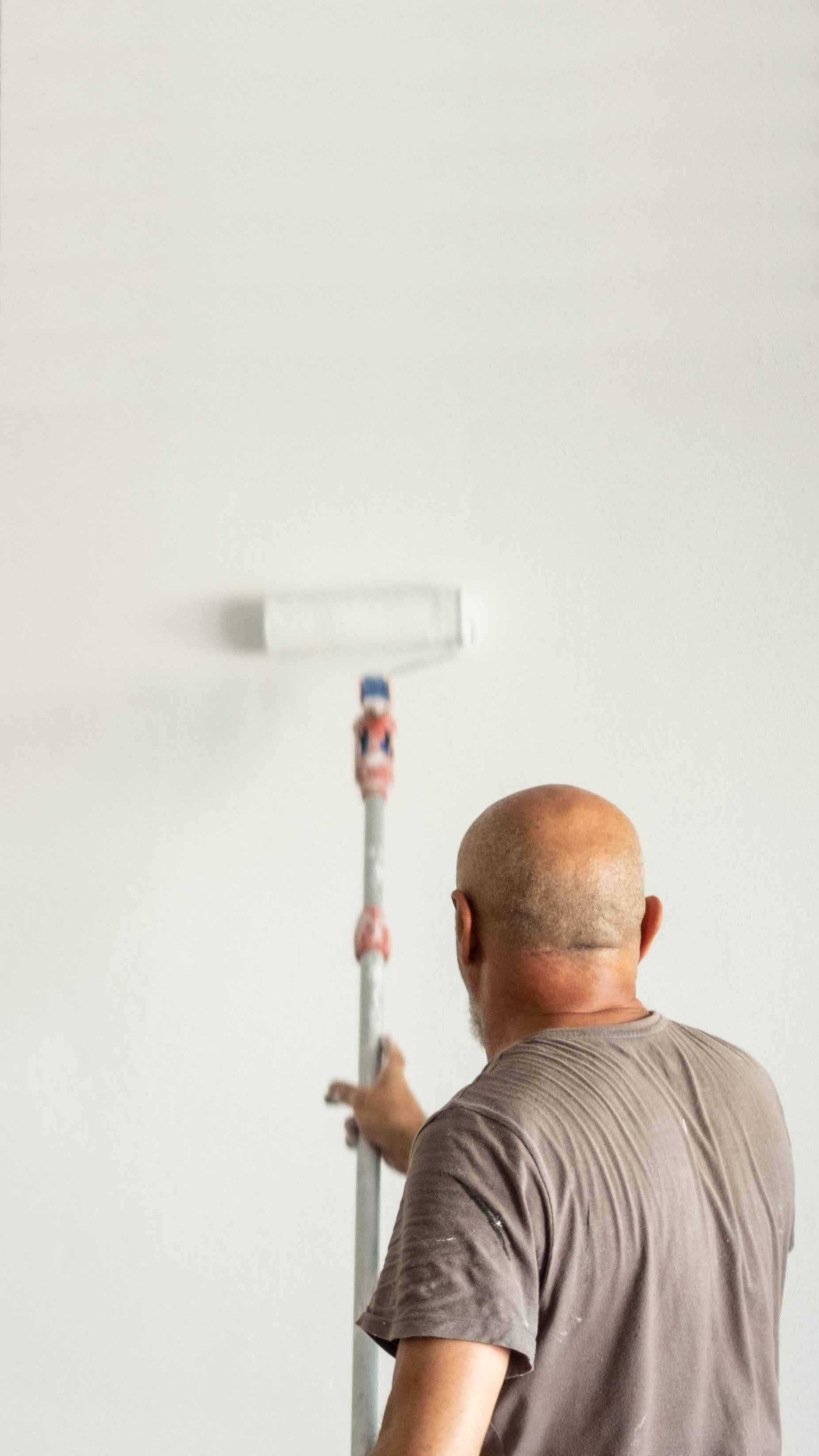 A person with a bald head paints a white wall using a roller on an extended pole, wearing a gray t-shirt, in a minimalistic setting.