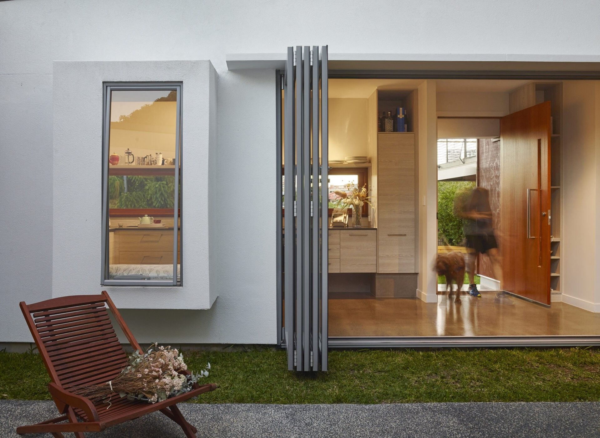 Modern home exterior with a large sliding door open to the kitchen. A person and a dog are near the entrance. A wooden chair with flowers is outside.