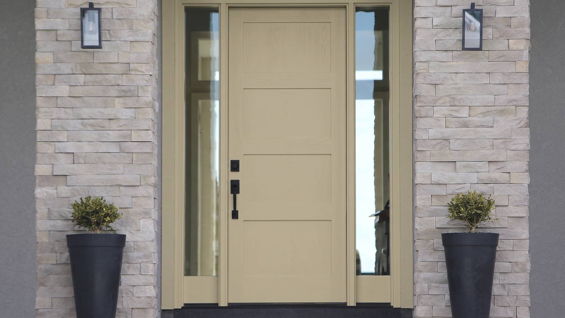 A wooden front door with sidelights surrounded by light stone bricks, flanked by two tall black planters with small shrubs.
