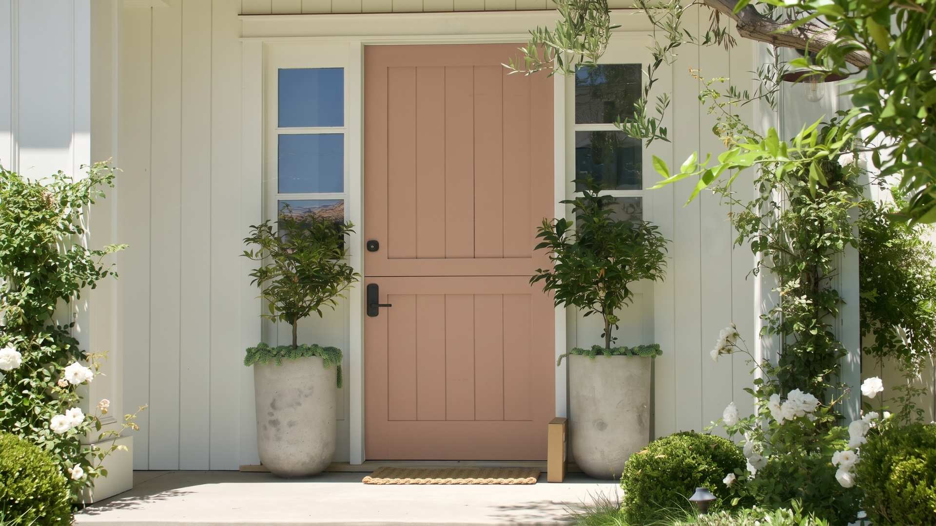 Cinnamon Doughnut front door with windows on either side, framed by white siding and flanked by potted plants and blooming white flowers in a garden setting.