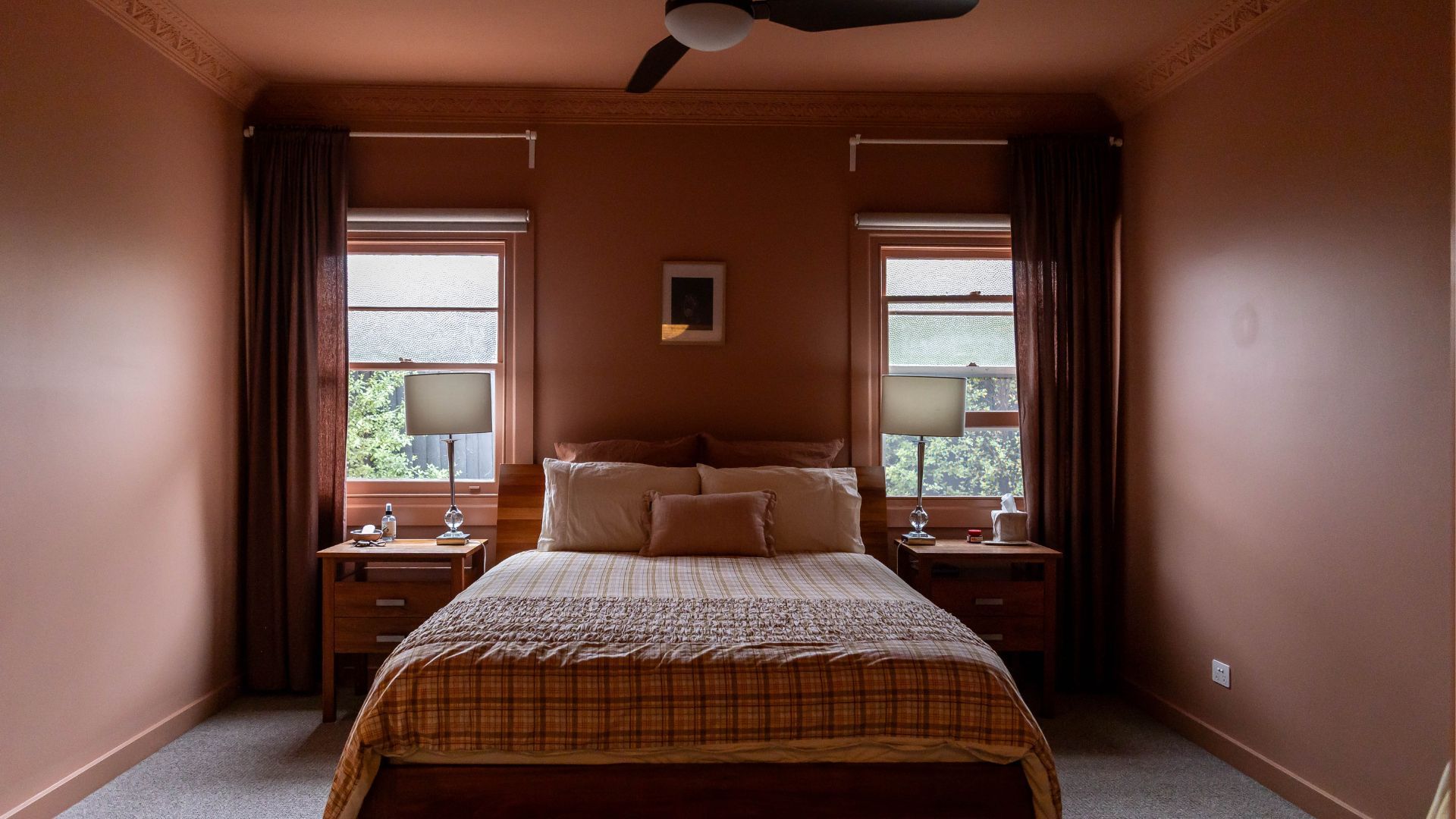 Cozy bedroom with brown walls, double bed, plaid bedspread, and two windows. Matching lamps and side tables flank the bed. Ceiling fan above, blush pink wall colour of Taubmans Pale Russet