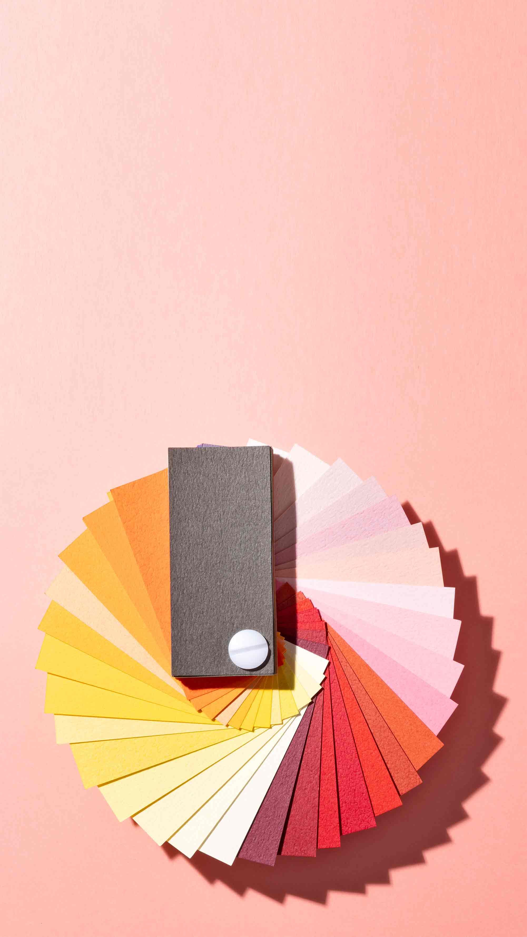 A spiral fan of colorful paper swatches on a pink background, featuring shades from red to yellow, with a gray swatch on top.