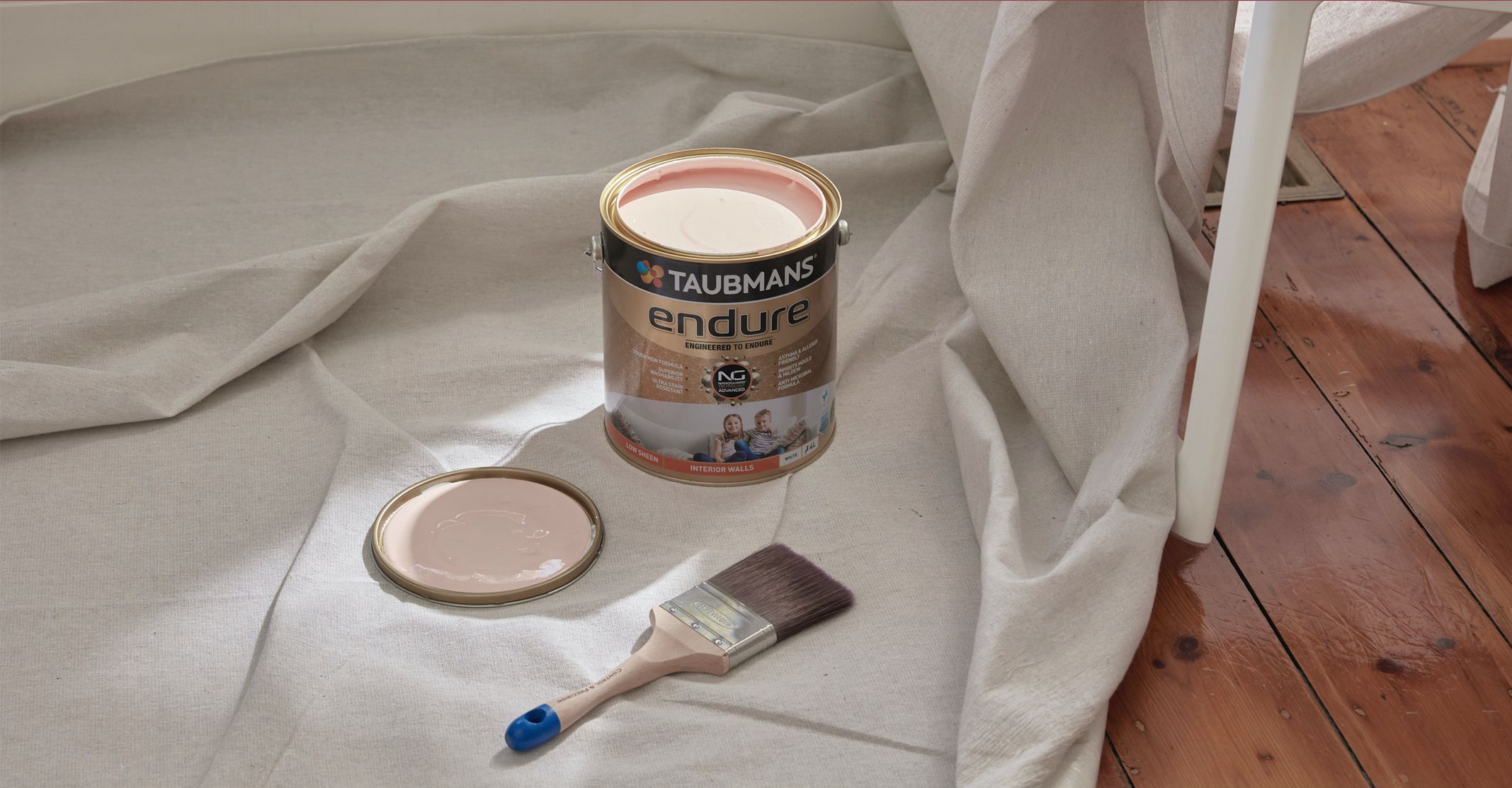 Paint can of Taubmans Endure with pink paint, lid, and brush on a drop cloth over wooden floor.