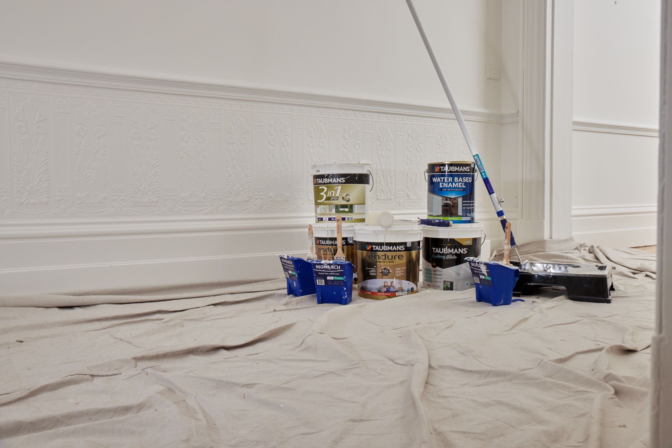 Buckets of paint and painting supplies are placed on a drop cloth in a room with ornate white walls.