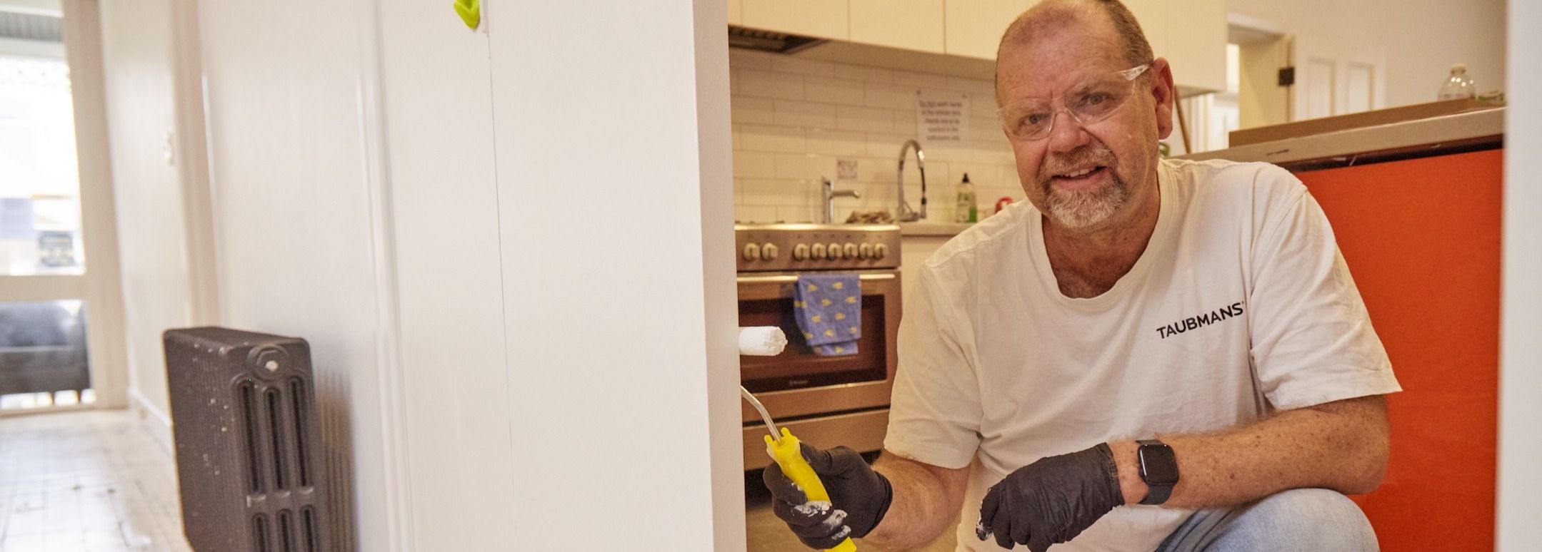 A man in a white shirt and gloves is painting a wall with a small roller in a kitchen. He is smiling and wearing safety glasses.