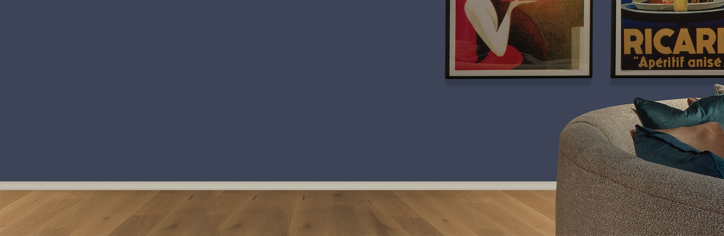 A cozy living room corner with a curved gray sofa, blue and brown cushions, wooden floor, and framed art on a dark blue wall.