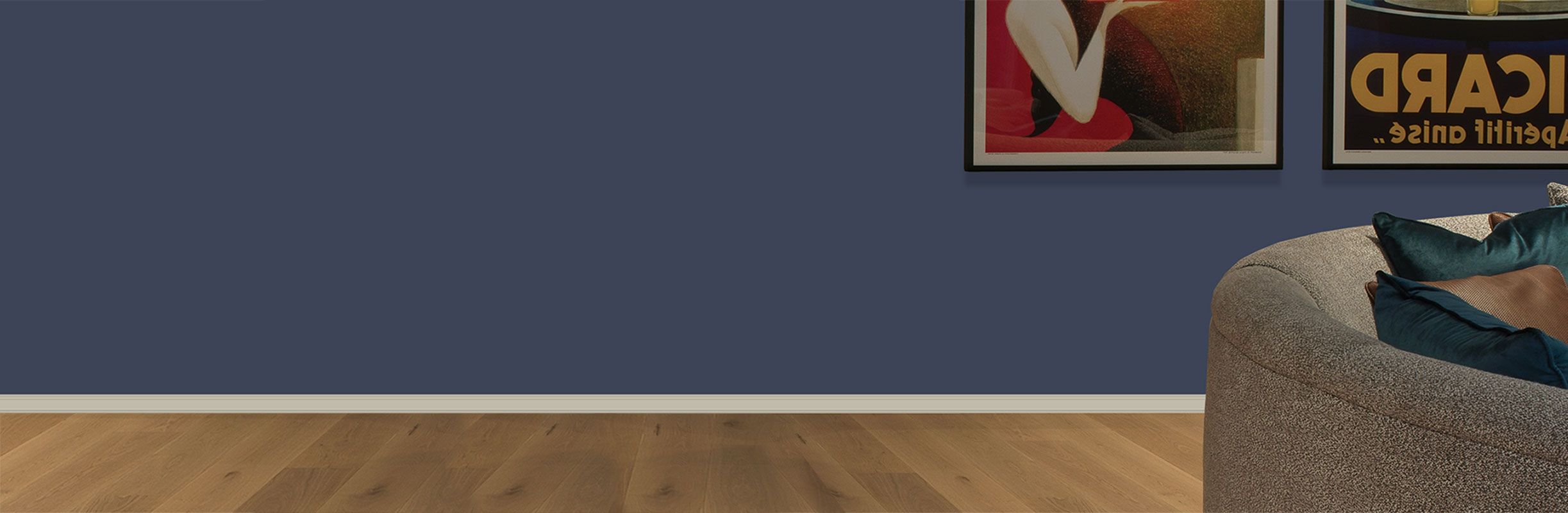 A cozy living room corner with a curved gray sofa, blue and brown cushions, wooden floor, and framed art on a dark blue wall.