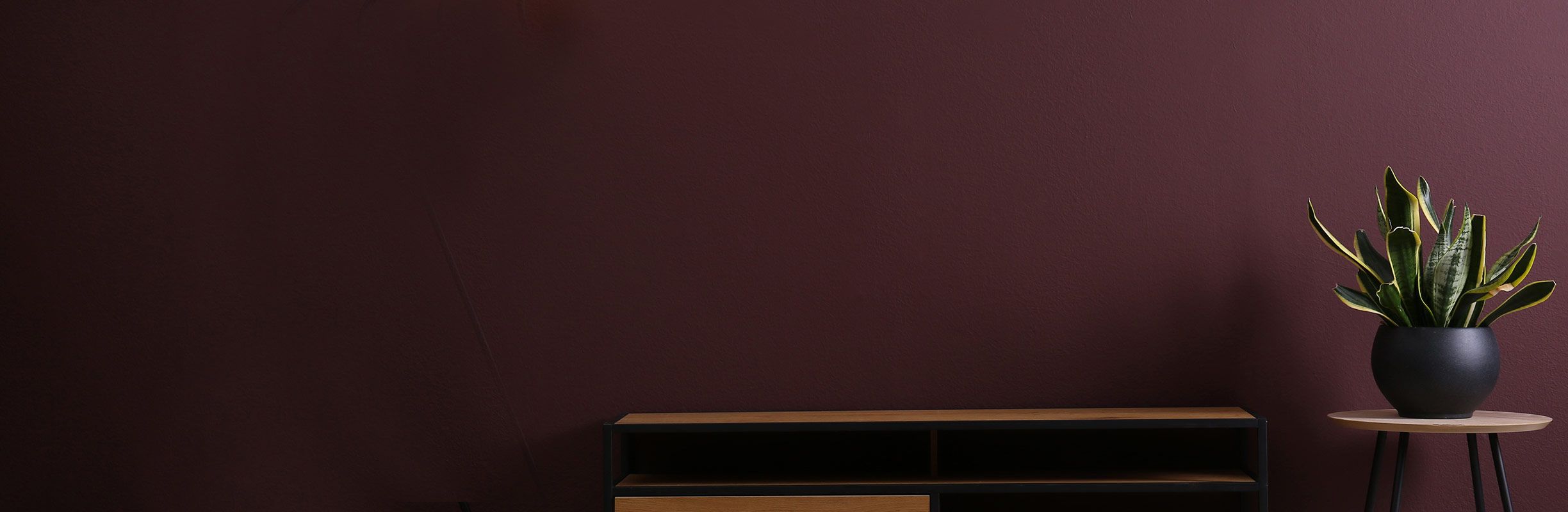 Minimalist room with a dark maroon wall, a black console with wooden accents, and a potted plant on a small round table.