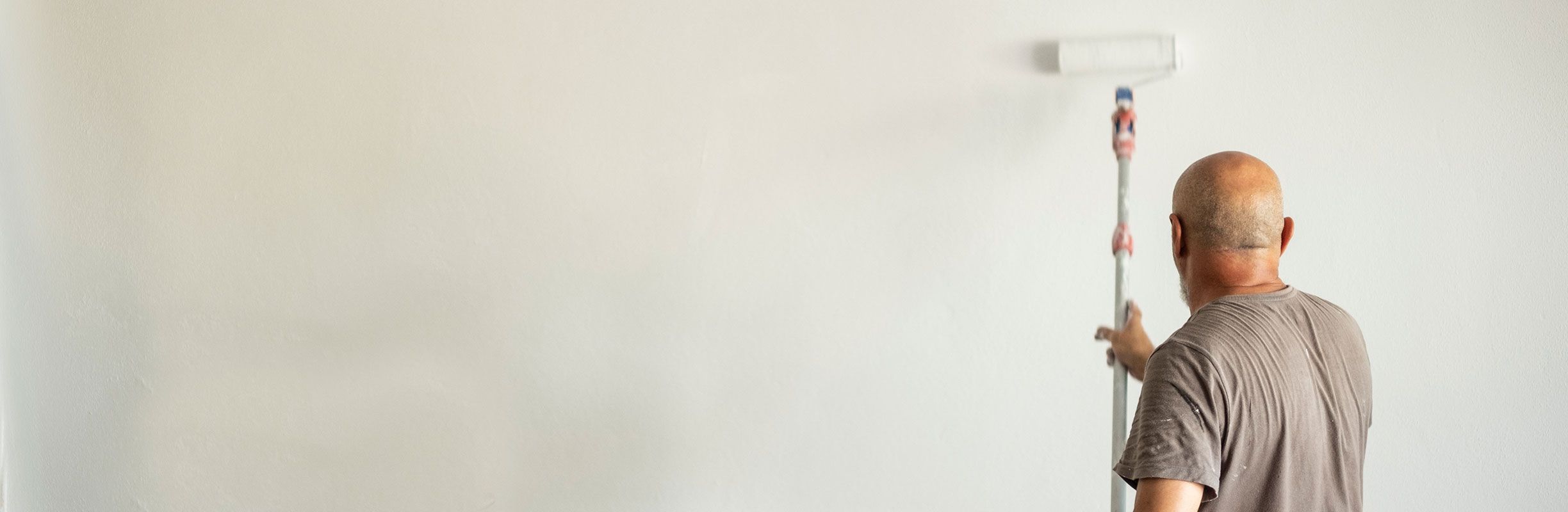 A person with a bald head paints a white wall using a roller on an extended pole, wearing a gray t-shirt, in a minimalistic setting.
