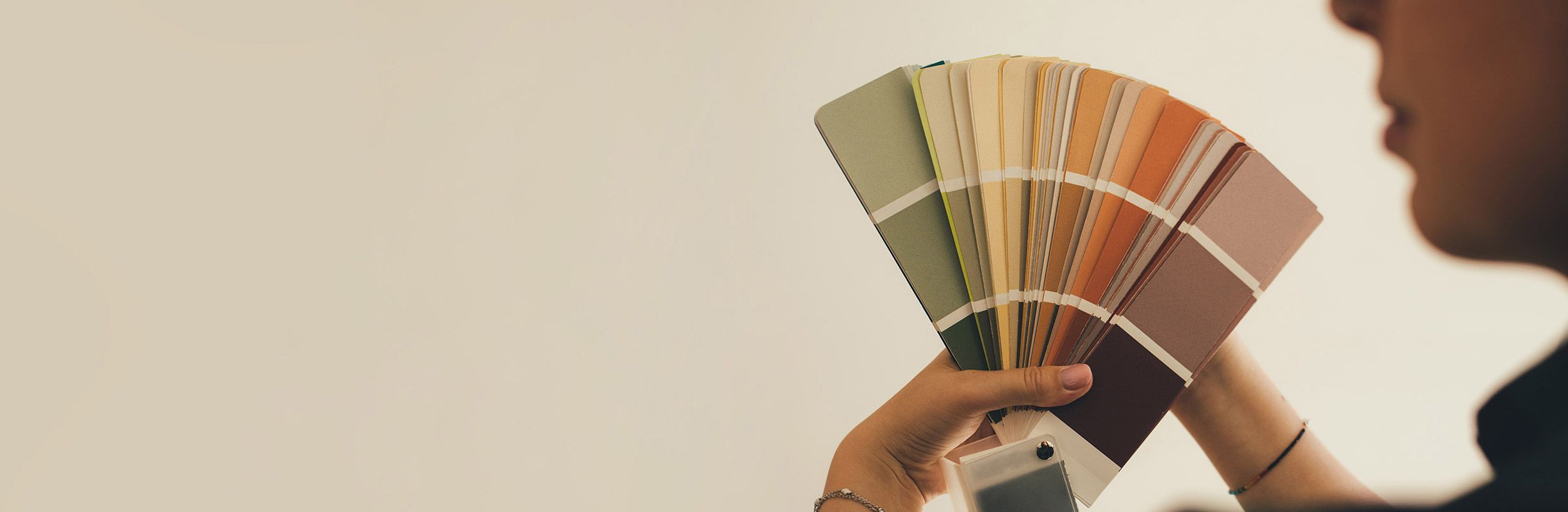 A person holding a fan of paint color swatches, featuring shades of green, yellow, orange, and brown, against a neutral background.