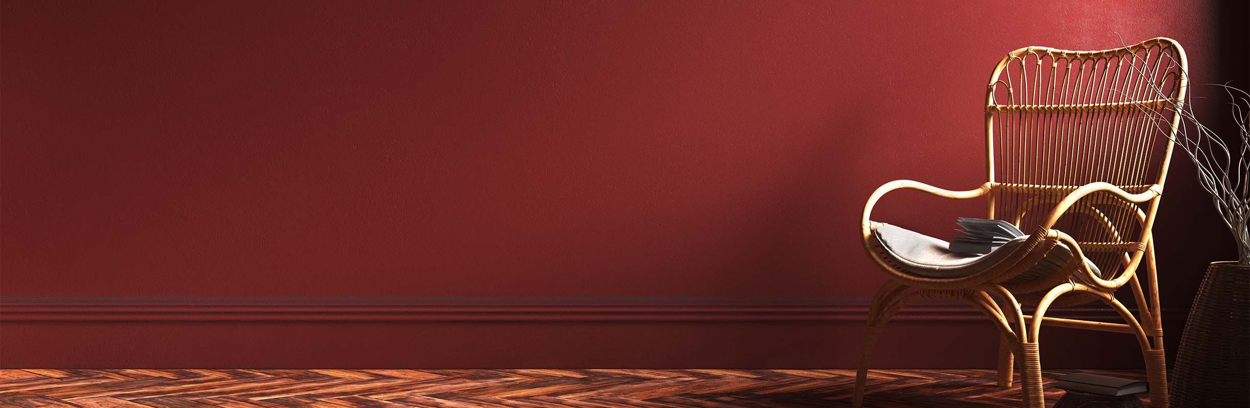 Wicker chair with a cushion and book on a wooden herringbone floor against a deep red wall, next to a vase with twigs.
