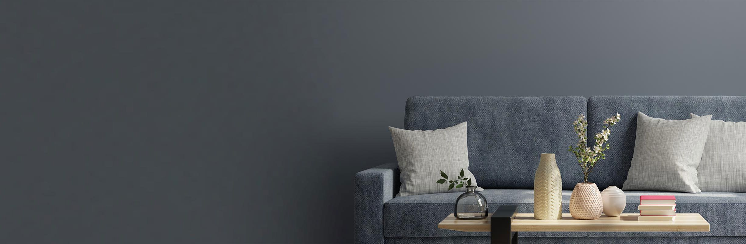A modern living room with a blue sofa, gray and white cushions, and a wooden table holding decorative vases and books against a dark wall.