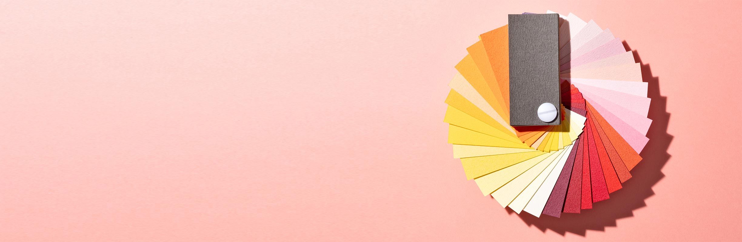 A spiral fan of colorful paper swatches on a pink background, featuring shades from red to yellow, with a gray swatch on top.