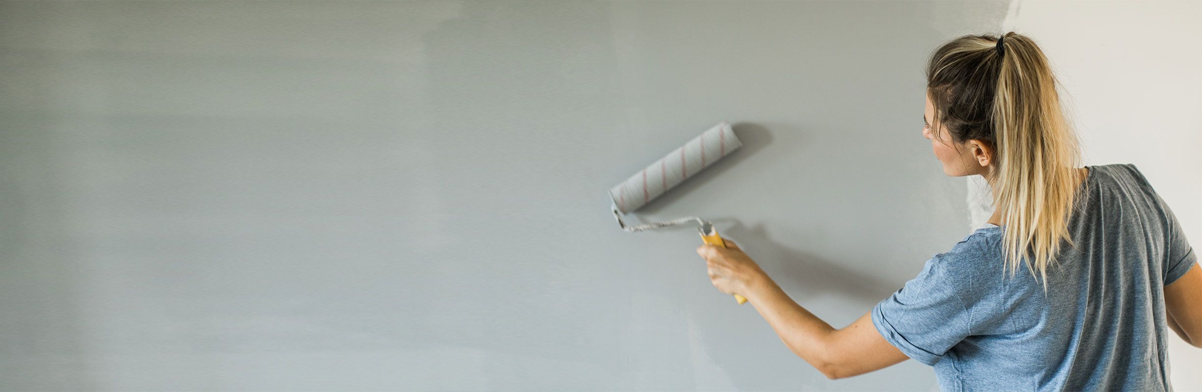 Woman painting a wall light gray with a roller. She wears a blue shirt and has her hair in a ponytail. The wall is partially painted.