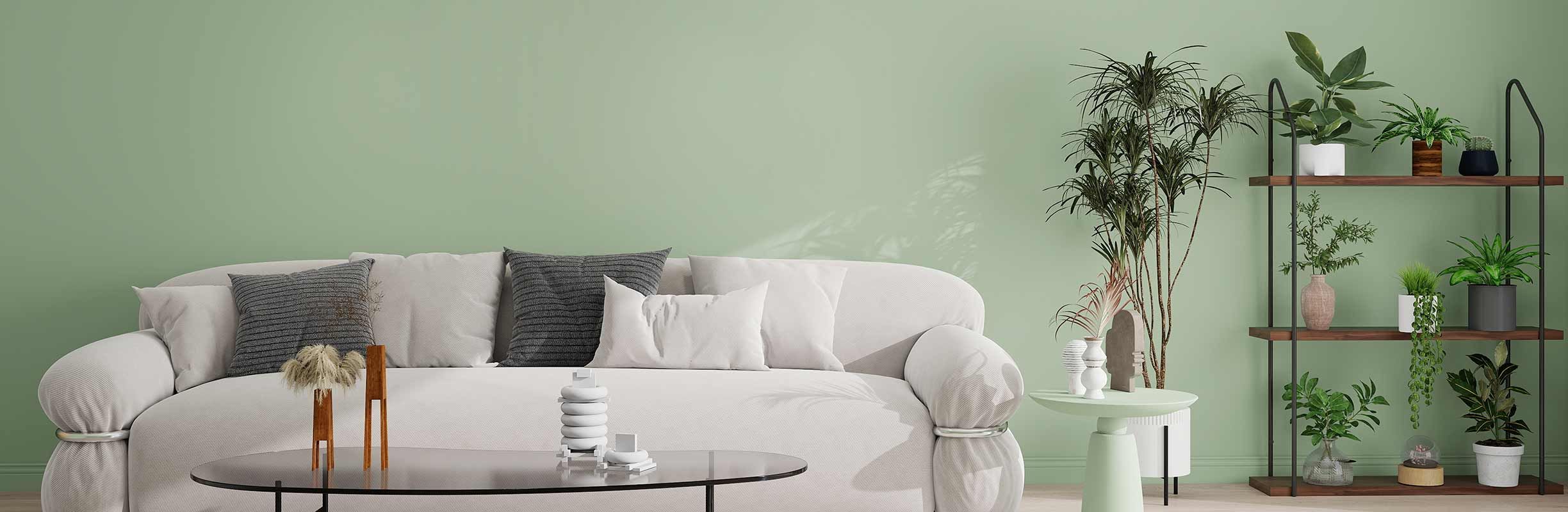A cozy living room with a white sofa, gray cushions, a round coffee table, and shelves of potted plants against a light green wall.