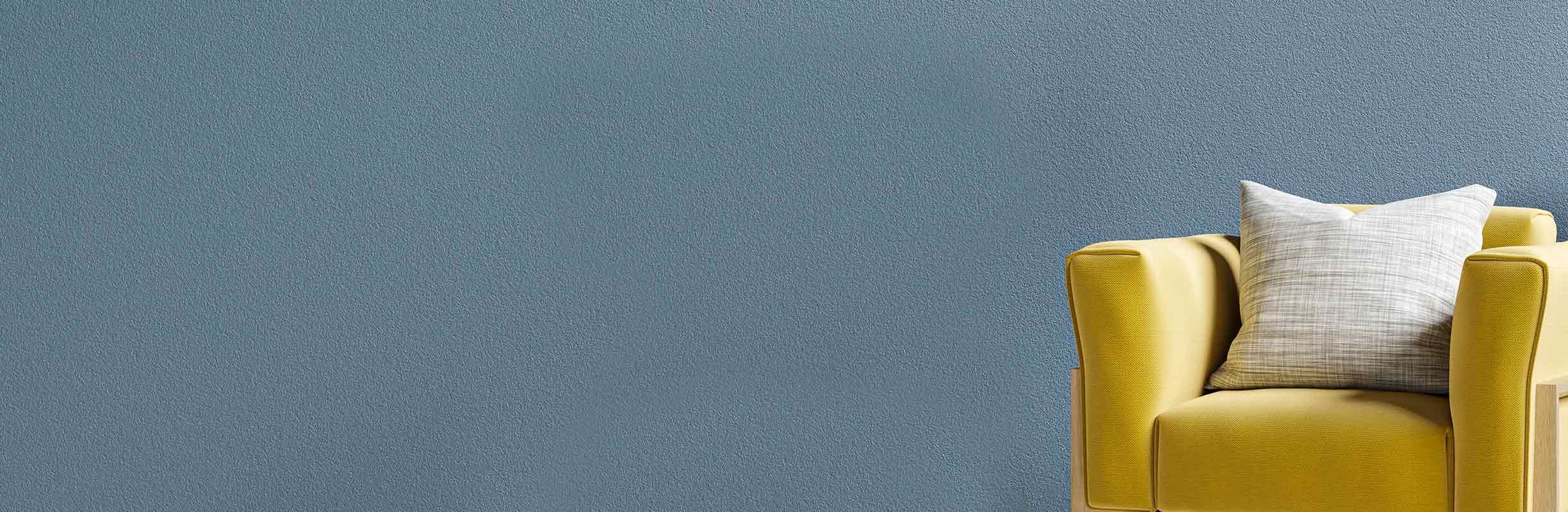 Yellow armchair with a light gray pillow against a textured blue-gray wall.
