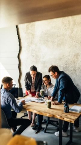 Four people in a meeting room gather around a wooden table, discussing documents. The room has a textured wall and warm lighting.