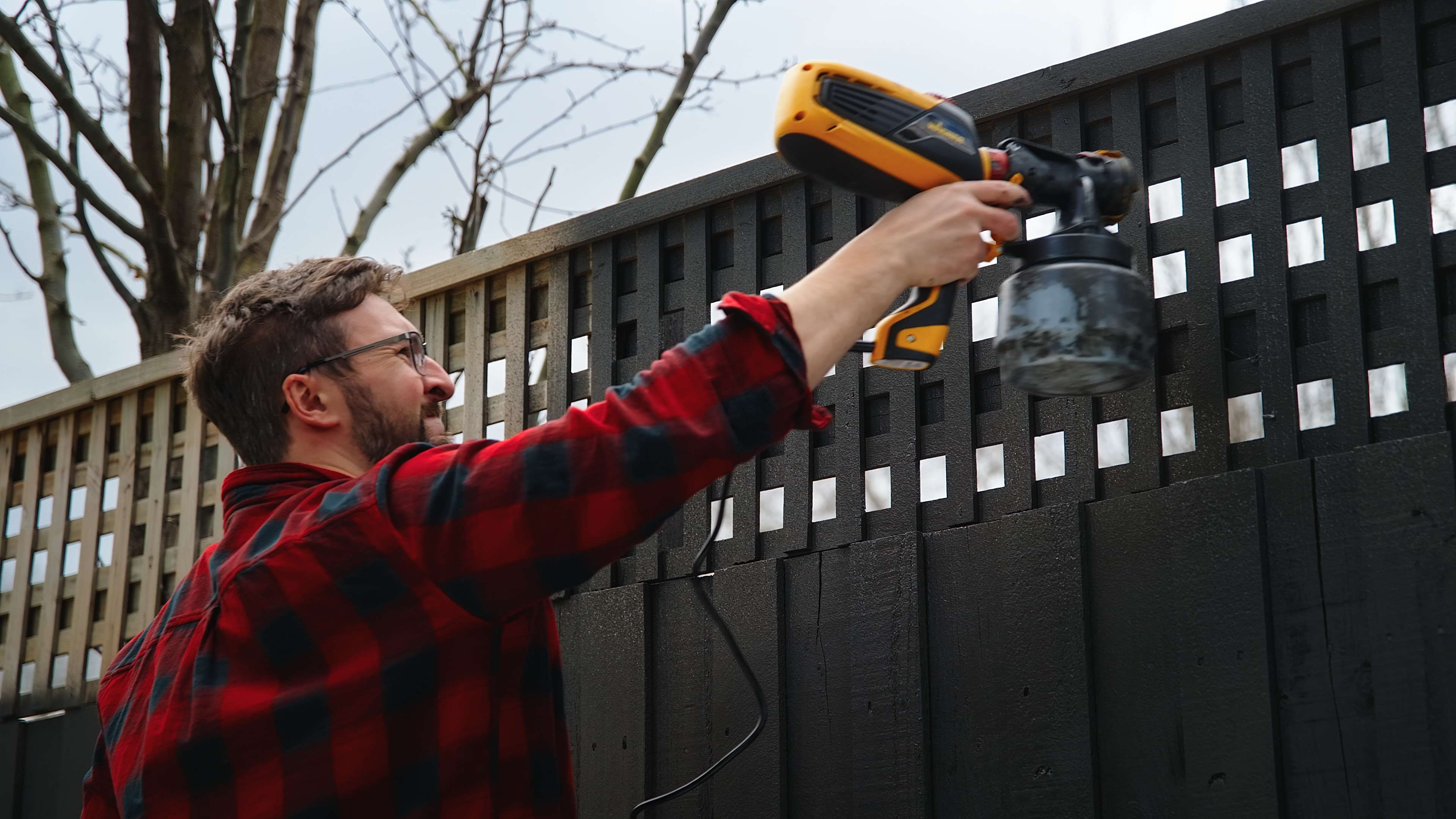 Man using spray gun to apply dark exterior paint onto fence