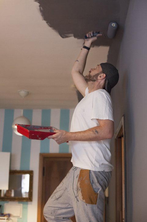 Man in casual clothes painting a ceiling with a roller, holding a red paint tray. Room has striped blue and white walls in the background.