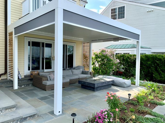 Modern patio with a white pergola, grey sectional sofa, and a rectangular fire pit. Surrounded by a garden and adjacent to a house.