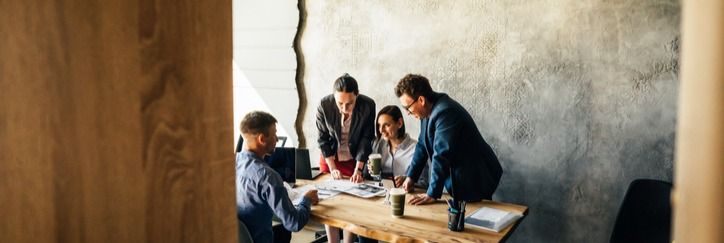 Four people in a meeting room gather around a wooden table, discussing documents. The room has a textured wall and warm lighting.